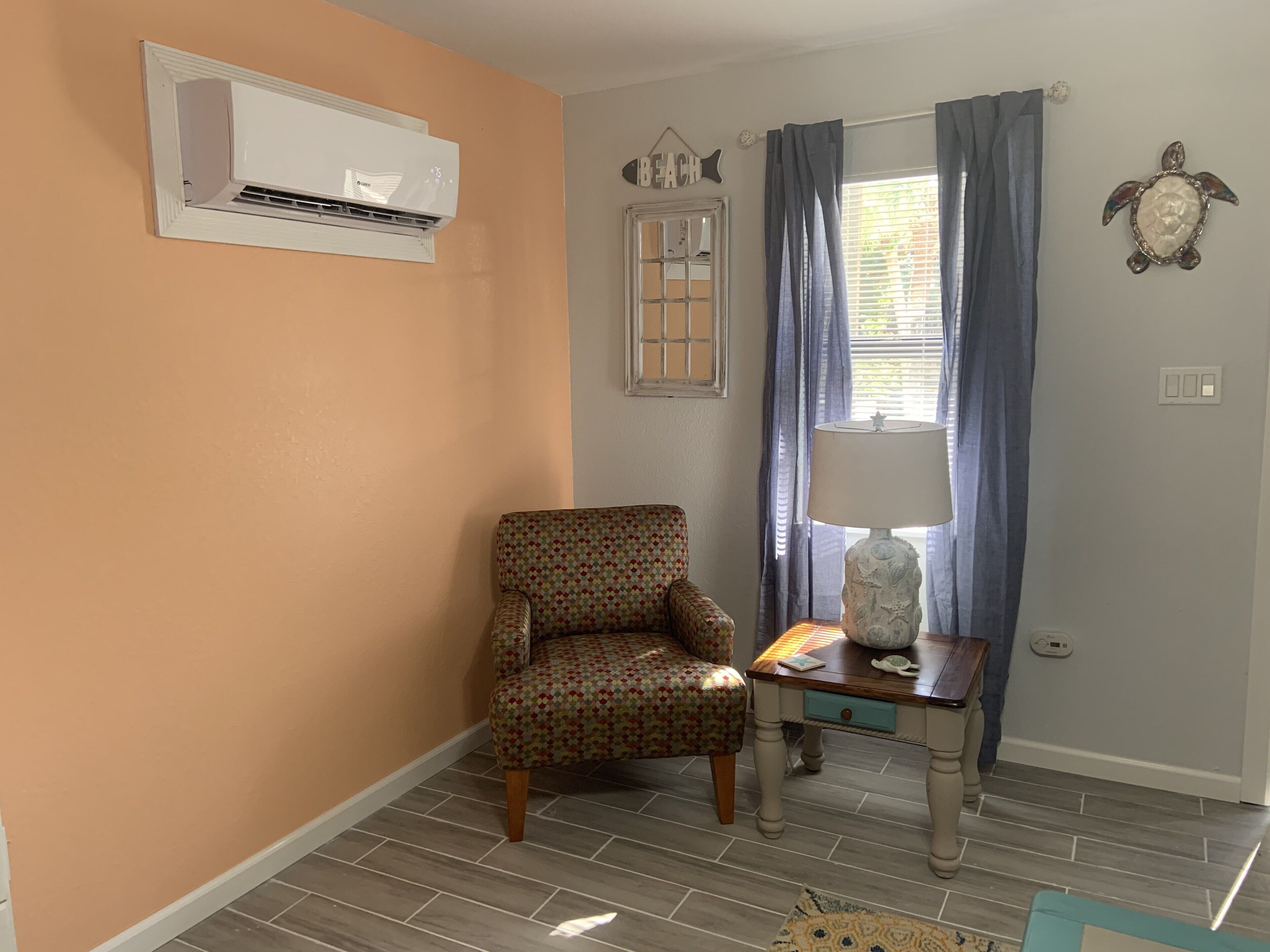 Une pièce comprenant un fauteuil à motifs, une petite table avec une lampe, un climatiseur fixé au mur et une fenêtre avec des rideaux.