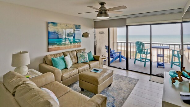 A living room with a beige sofa, a coffee table, and a painting of a beach scene. There is a balcony with blue chairs and a view of the ocean. - Modern Oceanfront condo for your Beach Getaway! Spectacular views! (South Padre Island)