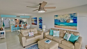 A spacious living room with a beige sofa set, a coffee table, and a ceiling fan. There is a kitchen area in the background with a dining set. - Modern Oceanfront condo for your Beach Getaway! Spectacular views! (South Padre Island)