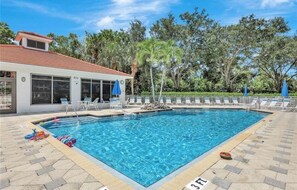 A swimming pool with lounge chairs and umbrellas, surrounded by trees and a building with large windows.