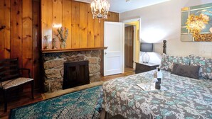 Ein Schlafzimmer mit Kamin aus Stein, Holzvertäfelung, einer Chandelierleuchtre, einem Bett mit gemusterten Betttextilien, einem Nachttisch und einem Sessel.
