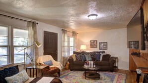 Ein gemütliches Wohnzimmer mit Sofa, Sessel und Couchtisch. Eine Tür, eine Lampe und ein Bücherregal sind ebenfalls vorhanden.