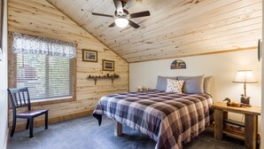 Un dormitorio acogedor con techo de madera, una cama con colcha a cuadros, una silla, una mesita de noche con lámpara y una ventana con persianas.