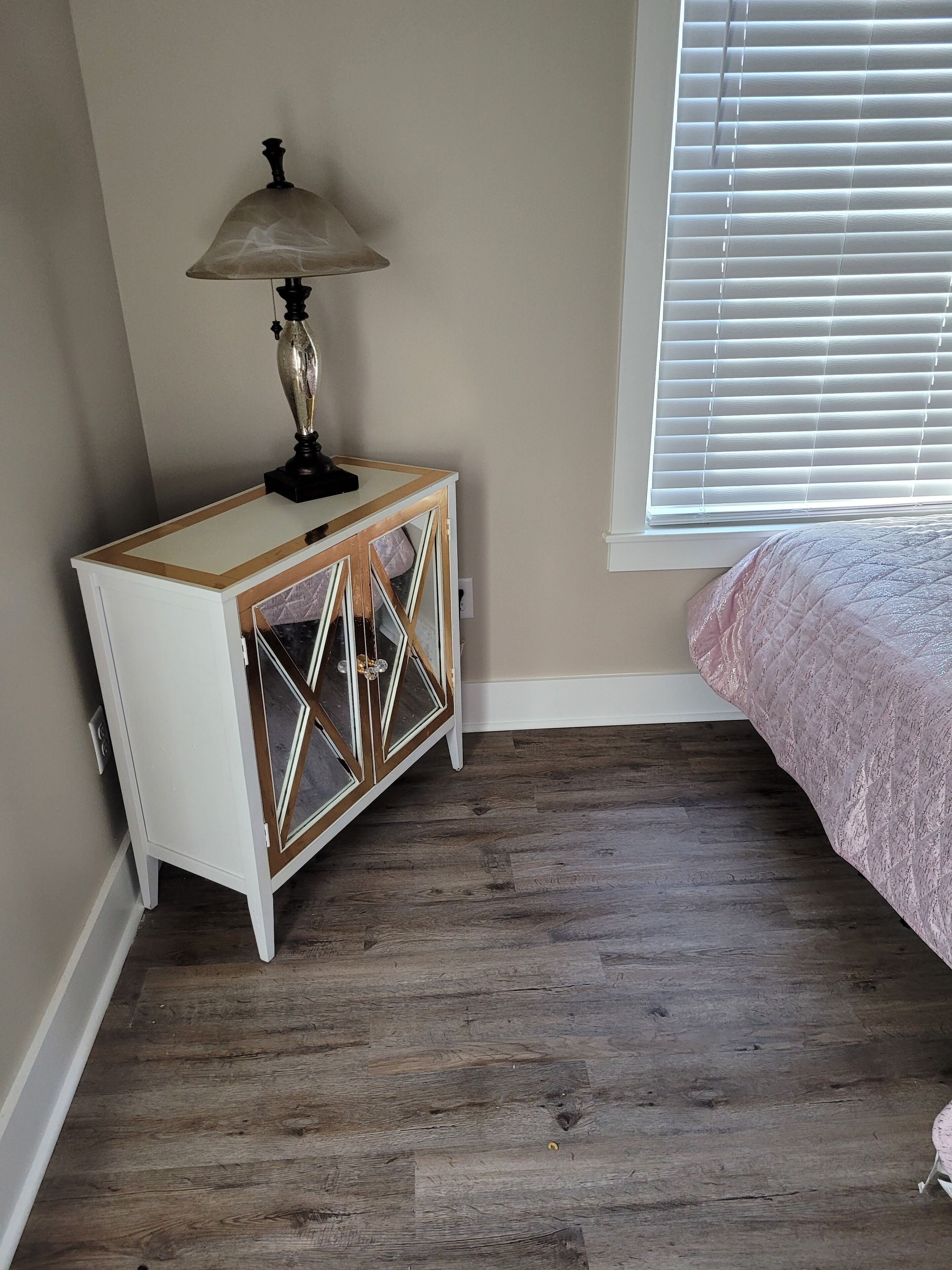 A bedroom with a wooden floor, a white bedside table with a lamp, and a window with blinds.