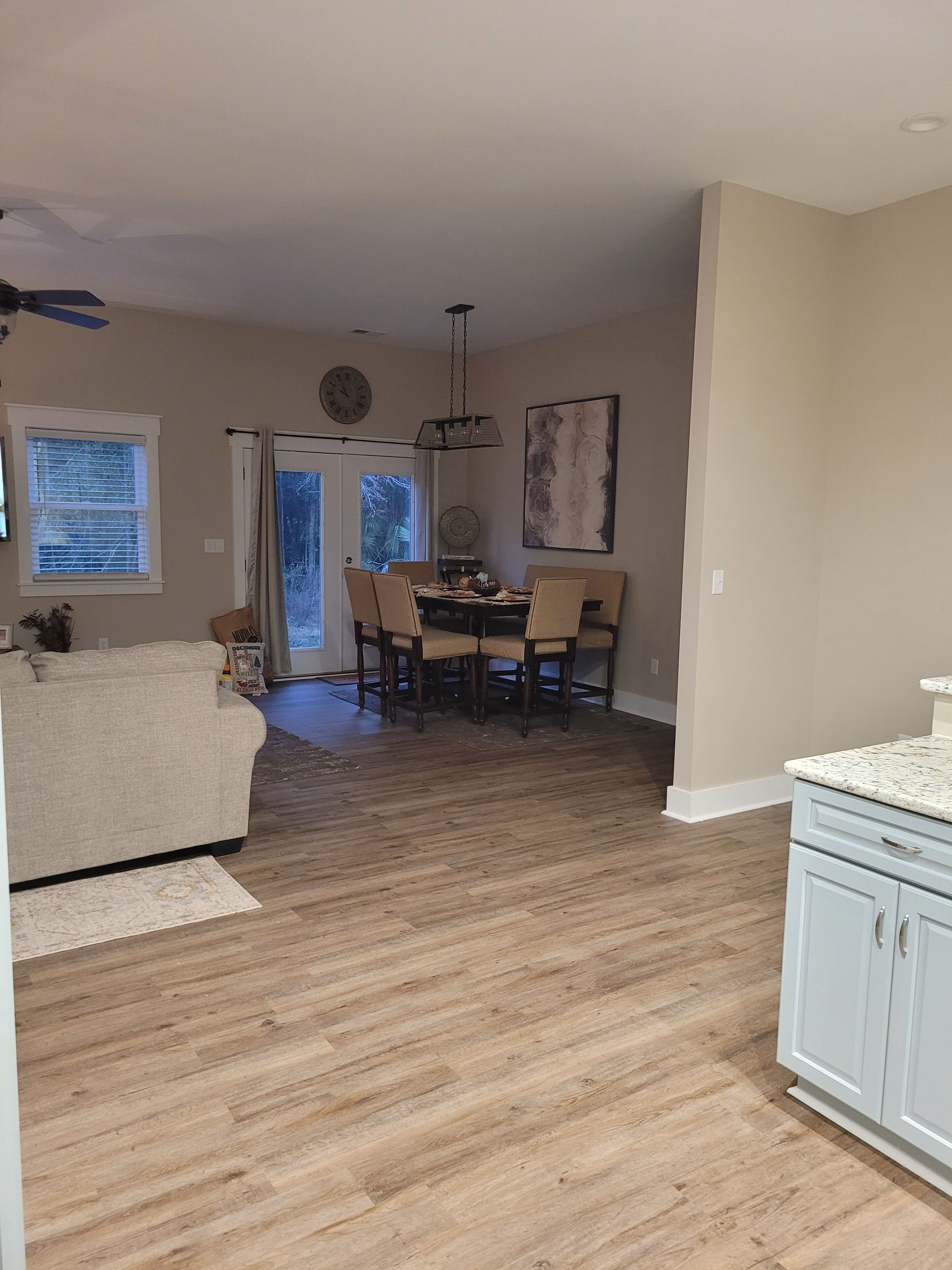 A spacious living room with a beige sofa, a dining area with a table and chairs, and a kitchen with white cabinets.