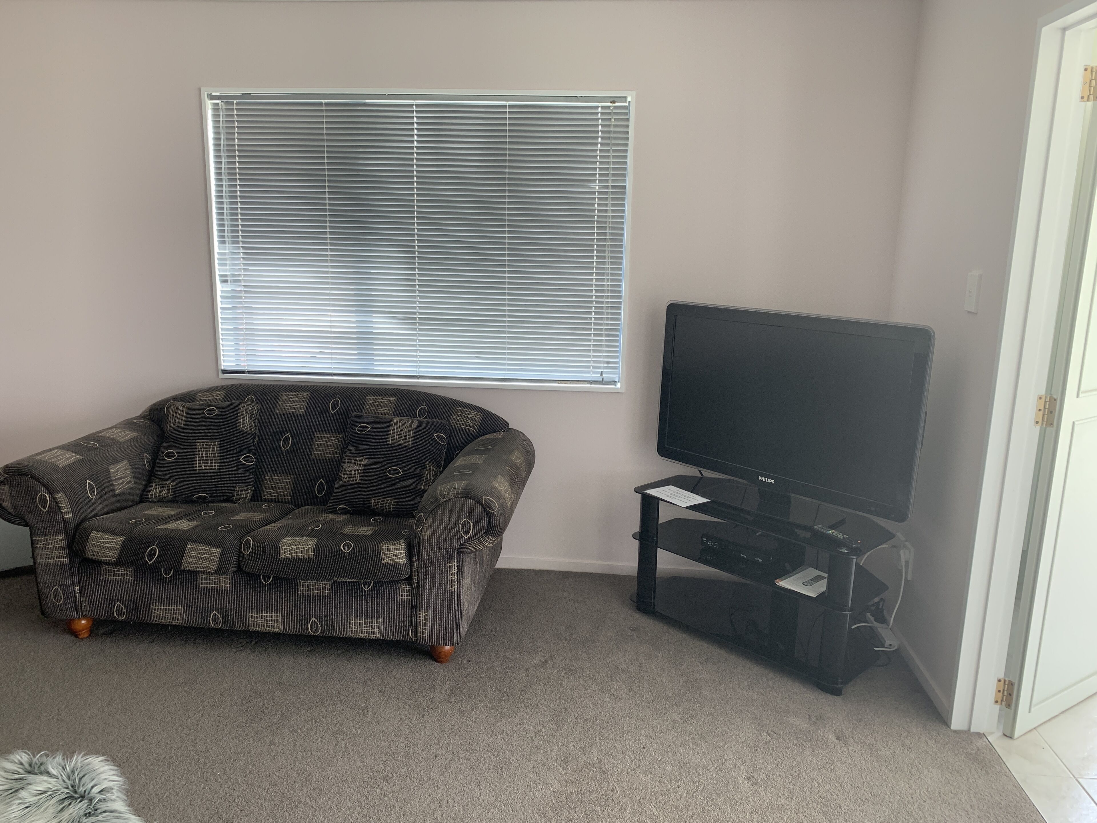 A living room with a patterned sofa, a television on a stand, and a window with blinds.