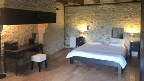 A rustic bedroom with exposed stone walls, a wooden ceiling, a bed with white linens, a desk with a chair, and two bedside tables with lamps. - Character house with swimming pool between Lot and Dordogne (Gourdon)