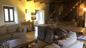 A cozy living room with a sofa, a coffee table, and wicker chairs. There is a dining table with a vase of flowers. The room has wooden beams on the ceiling and a staircase in the background. - Character house with swimming pool between Lot and Dordogne (Gourdon)