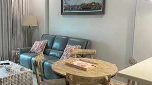 Ein modernes Wohnzimmer mit einem blauen Ledersofa, einem Holzesstisch mit Stühlen, einem gemusterten Hocker, einem Gemälde mit Küstenlandschaft und einer Stehlampe.