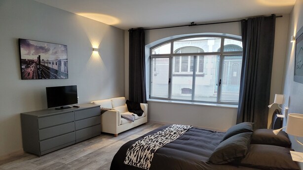 Une chambre moderne avec un grand lit, un téléviseur posé sur une commode, un canapé et une fenêtre avec des rideaux.