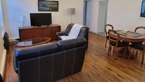 Un salon comprenant un canapé en cuir, une table basse en bois, un téléviseur à écran plat posé sur un meuble en bois, une table à manger avec des chaises et un tableau accroché au mur.