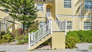 A multi-story building with a yellow exterior, white railings on the exterior staircases, and a well-maintained garden with various plants and a large tree. - Beachfront Resort Condo, Walk to Atlantis (Paradise Island)