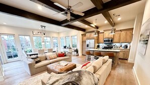 A spacious living room with a large beige sofa, a coffee table, and a fireplace. There is a kitchen area with wooden cabinets and a large island. The room has a high ceiling with exposed beams and a ceiling fan. - 15% off gorgeous cottage, hot tub, pool, fishing ponds, streams, waterfalls! (Vinita)