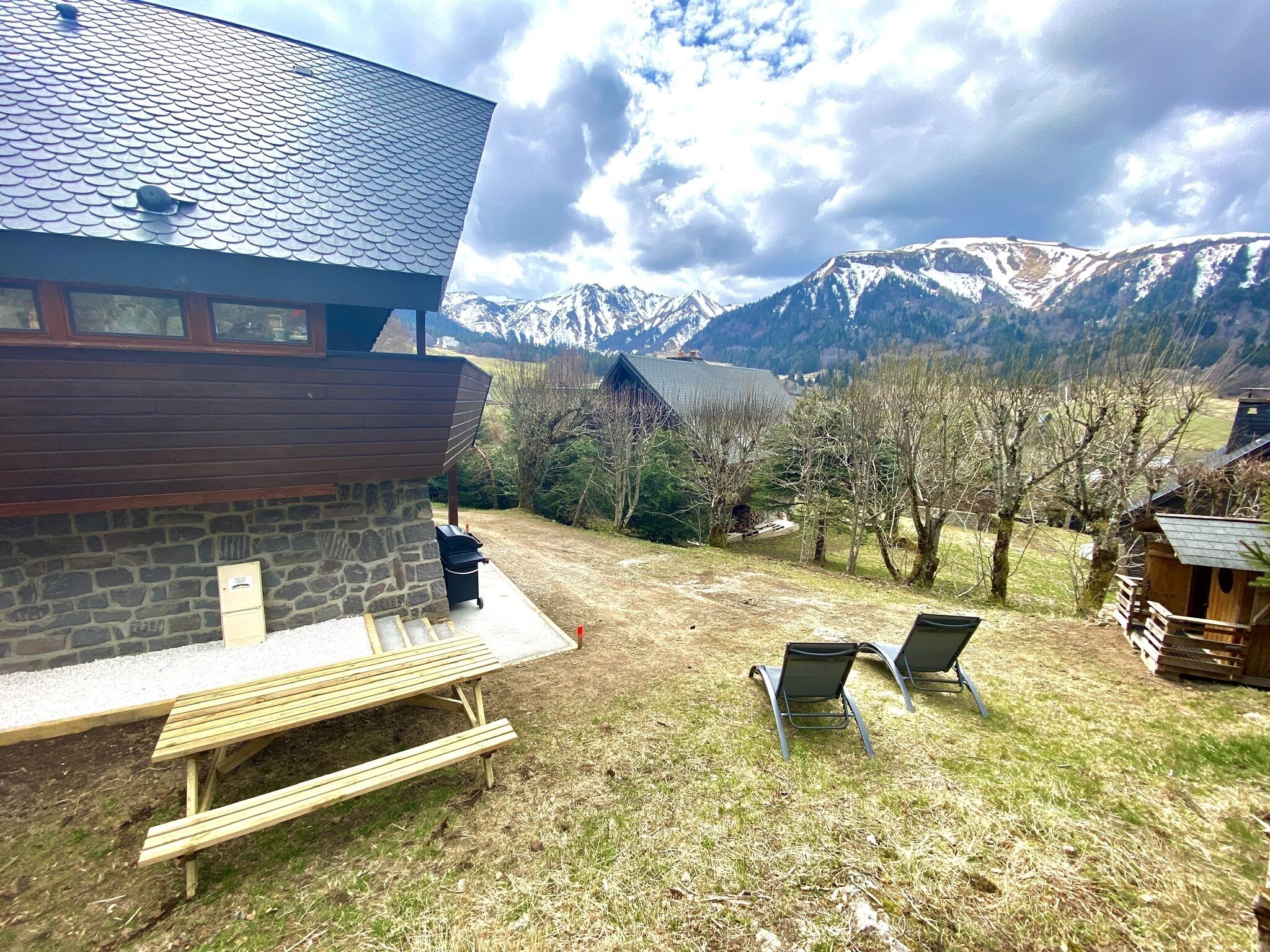 Un bâtiment en pierre avec un toit en pente, un banc en bois et deux chaises en métal, situés dans une cour verdoyante offrant une vue sur la montagne.