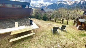 Un edificio de piedra con techo inclinado, un banco de madera y dos sillas de metal en un patio cubierto de césped con vista a la montaña.