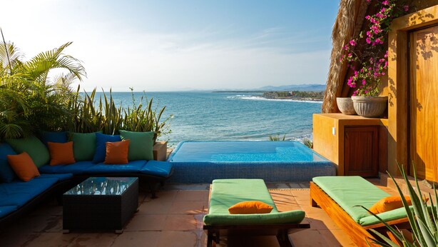 A poolside area with a view of the ocean, a wooden bench, a sofa with cushions, and a potted plant.