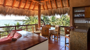 A room with a thatched roof, wooden furniture, a view of the ocean, and a bar area with shelves.