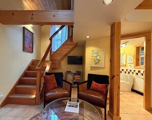 A spacious living area with a wooden staircase, leather armchairs, a glass coffee table with an informational placard, and a television mounted on the wall.