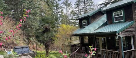 Eine Holzhütte mit Veranda, umgeben von einem Garten mit verschiedenen Pflanzen und einem großen Baum.