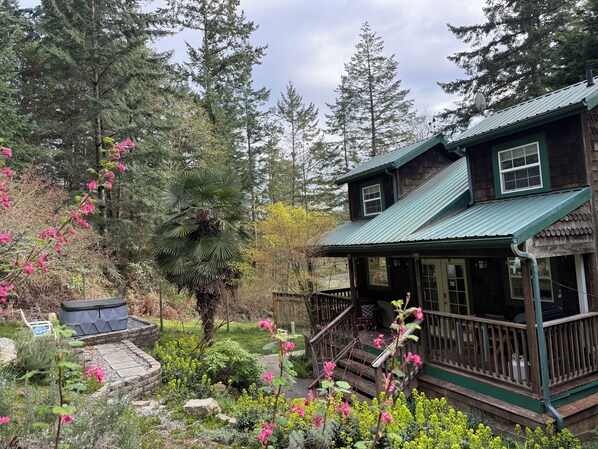 A wooden cabin with a porch, surrounded by a garden with various plants and a large tree.