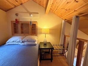 Un dormitorio con una cama, una mesa con una lámpara, una lámpara colgante y techo de madera.