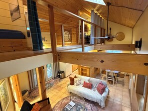Habitación estilo loft con techo alto, vigas de madera, una mesa de centro de vidrio, un sofá blanco con cojines estampados y vistas al exterior.