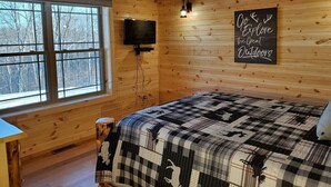 Une chambre chaleureuse dans un chalet, avec des murs en bois, une grande fenêtre, un lit recouvert d’une couverture à carreaux, un téléviseur fixé au mur et un panneau affichant un message motivant.
