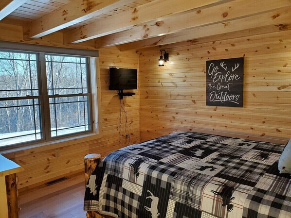 A cozy cabin room with wooden walls, a large window, a bed with a checkered blanket, a mounted television, and a motivational sign on the wall.