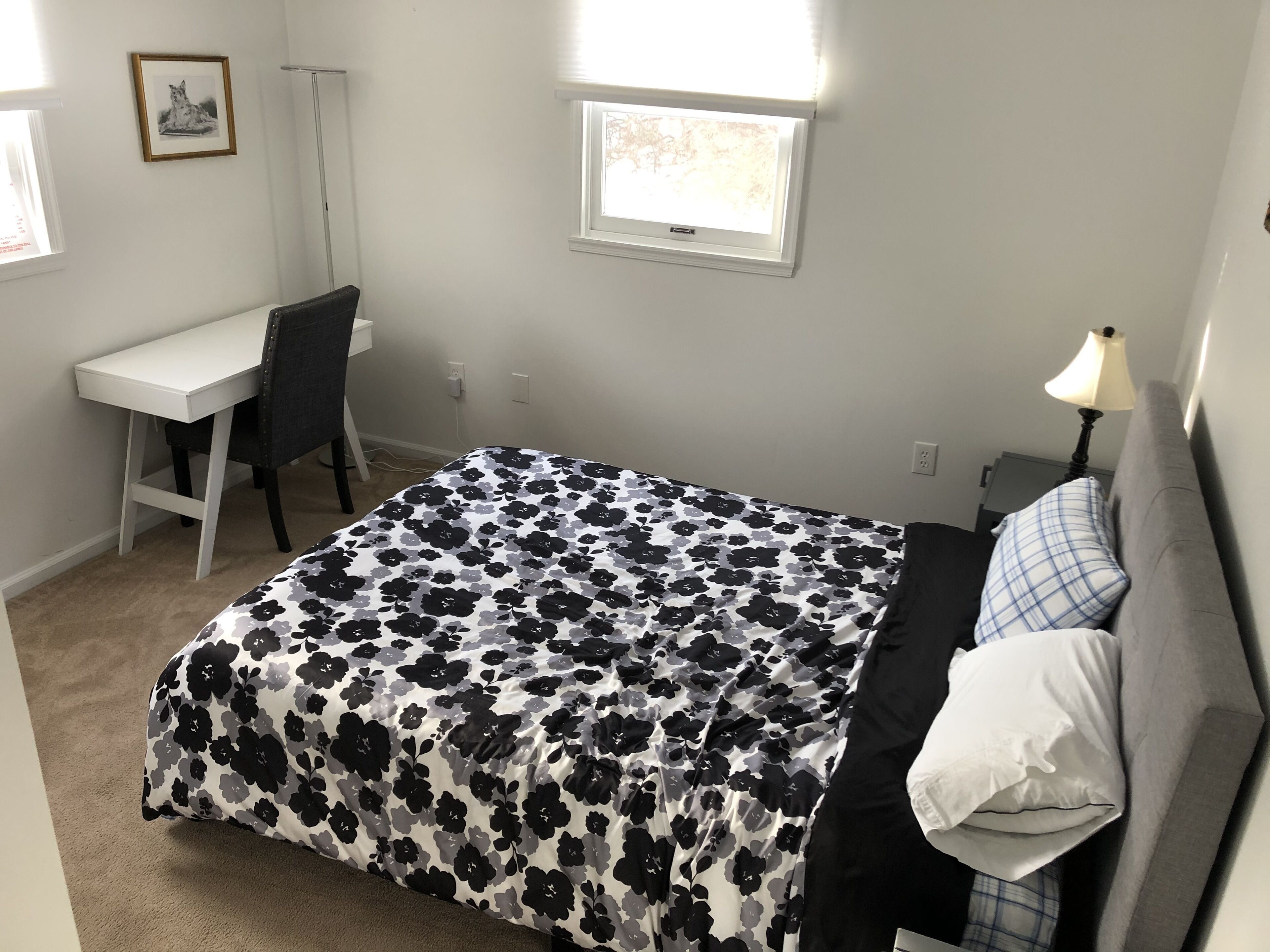 A bedroom with a bed, a desk, and a chair. There is a window with blinds, a lamp, and a framed picture on the wall.