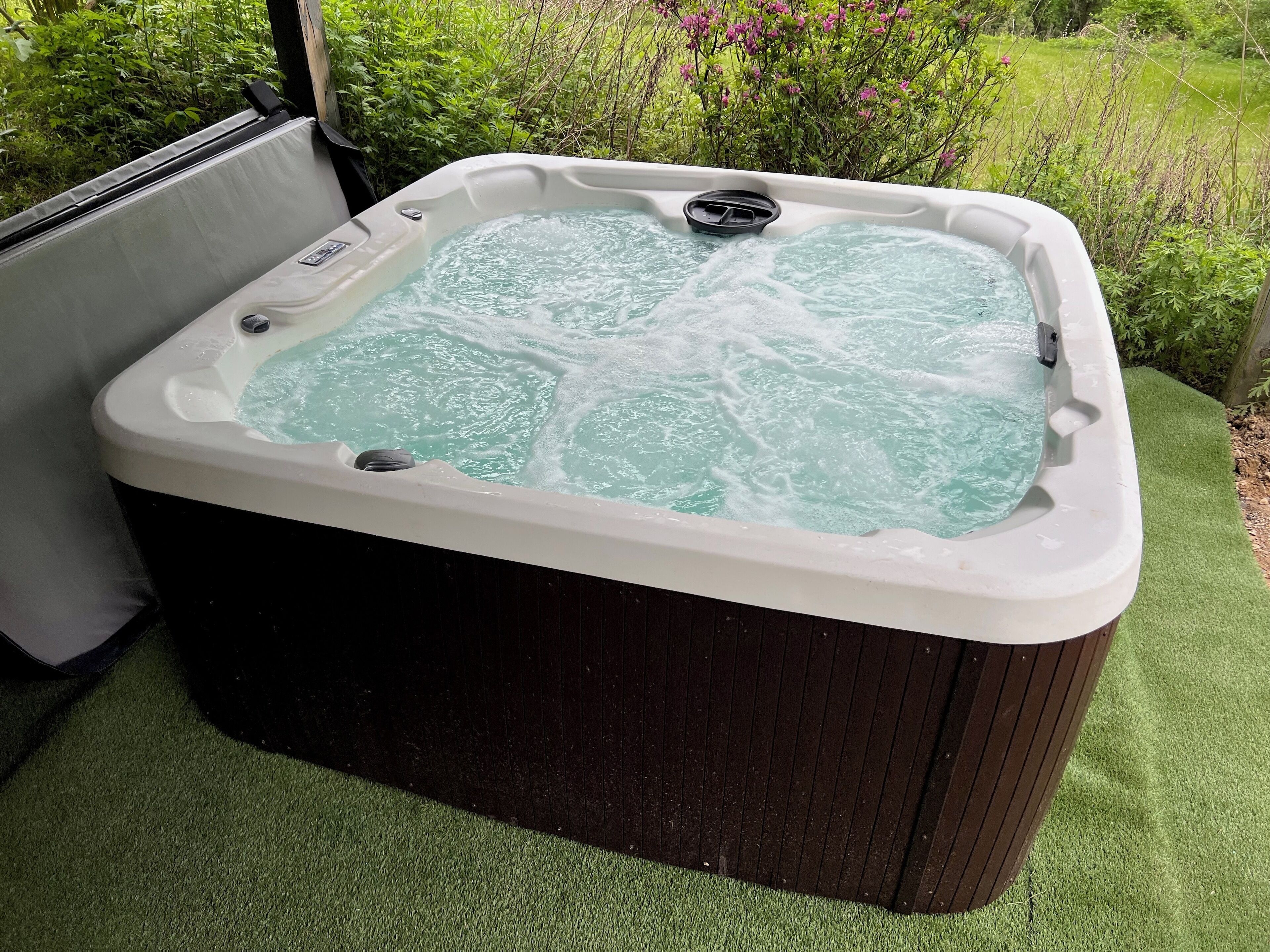A hot tub with a wooden exterior, surrounded by green artificial grass and lush vegetation.