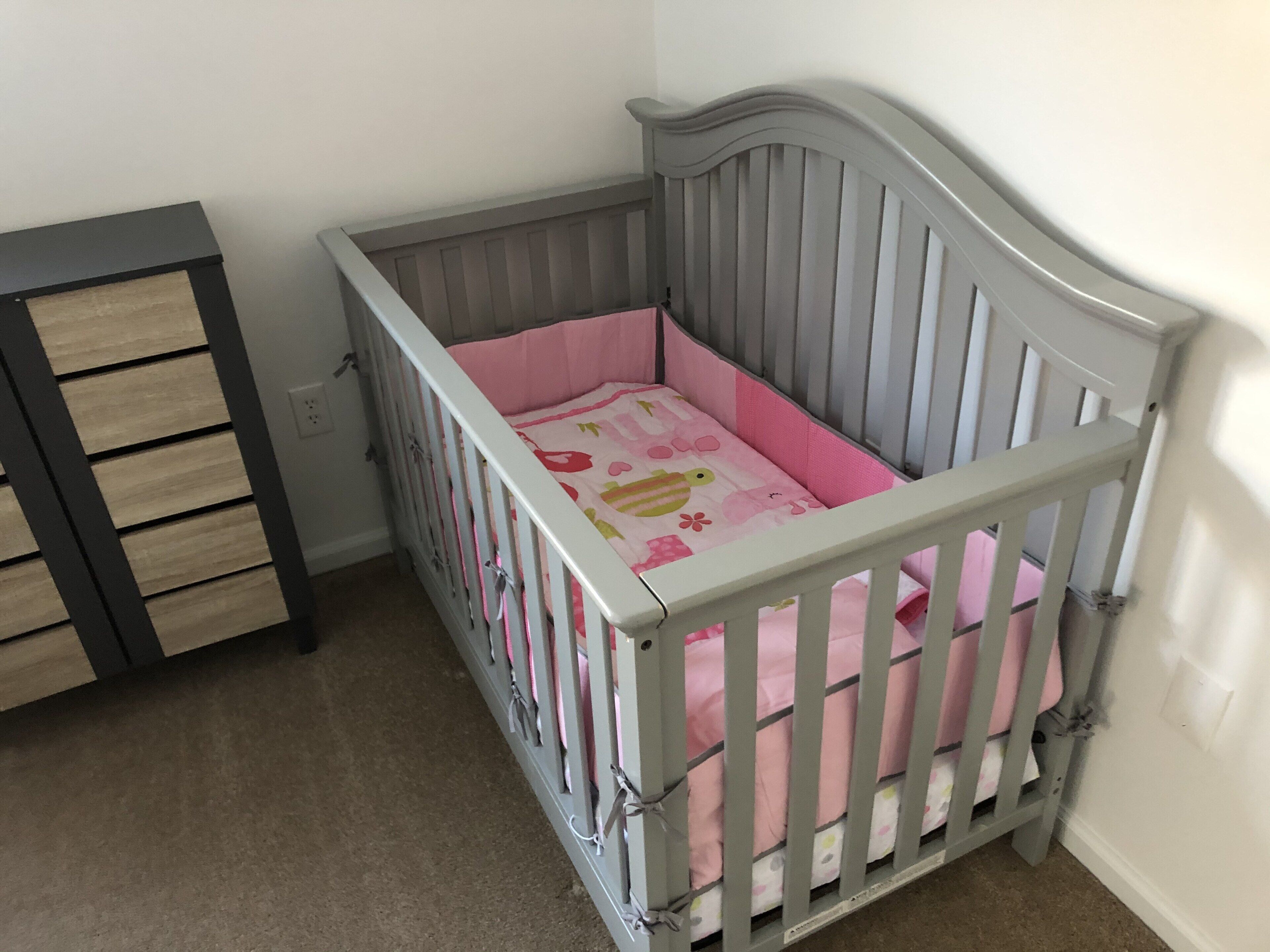 Una cuna para bebé con colchón rosa y cabecera gris, ubicada junto a un aparador de madera con varios cajones.