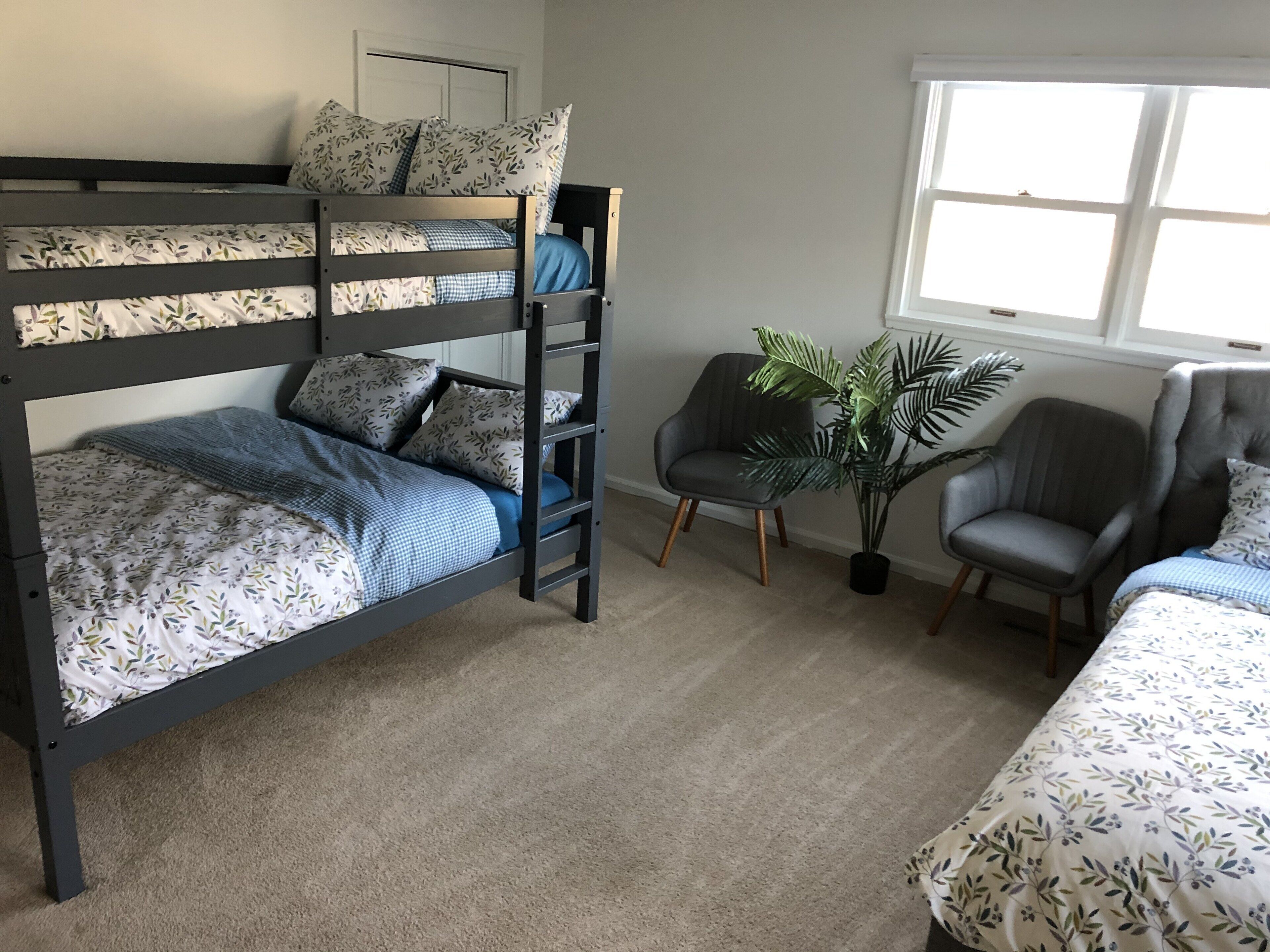 A bunk bed with floral bedding, a grey armchair, a potted plant, and a window with natural light.