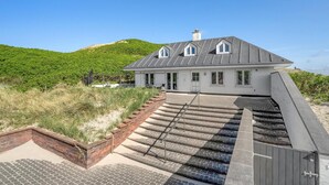 Une maison moderne avec un toit en pente, un escalier en briques menant à une entrée fermée par un portail, et une vue sur une colline verdoyante en arrière-plan.