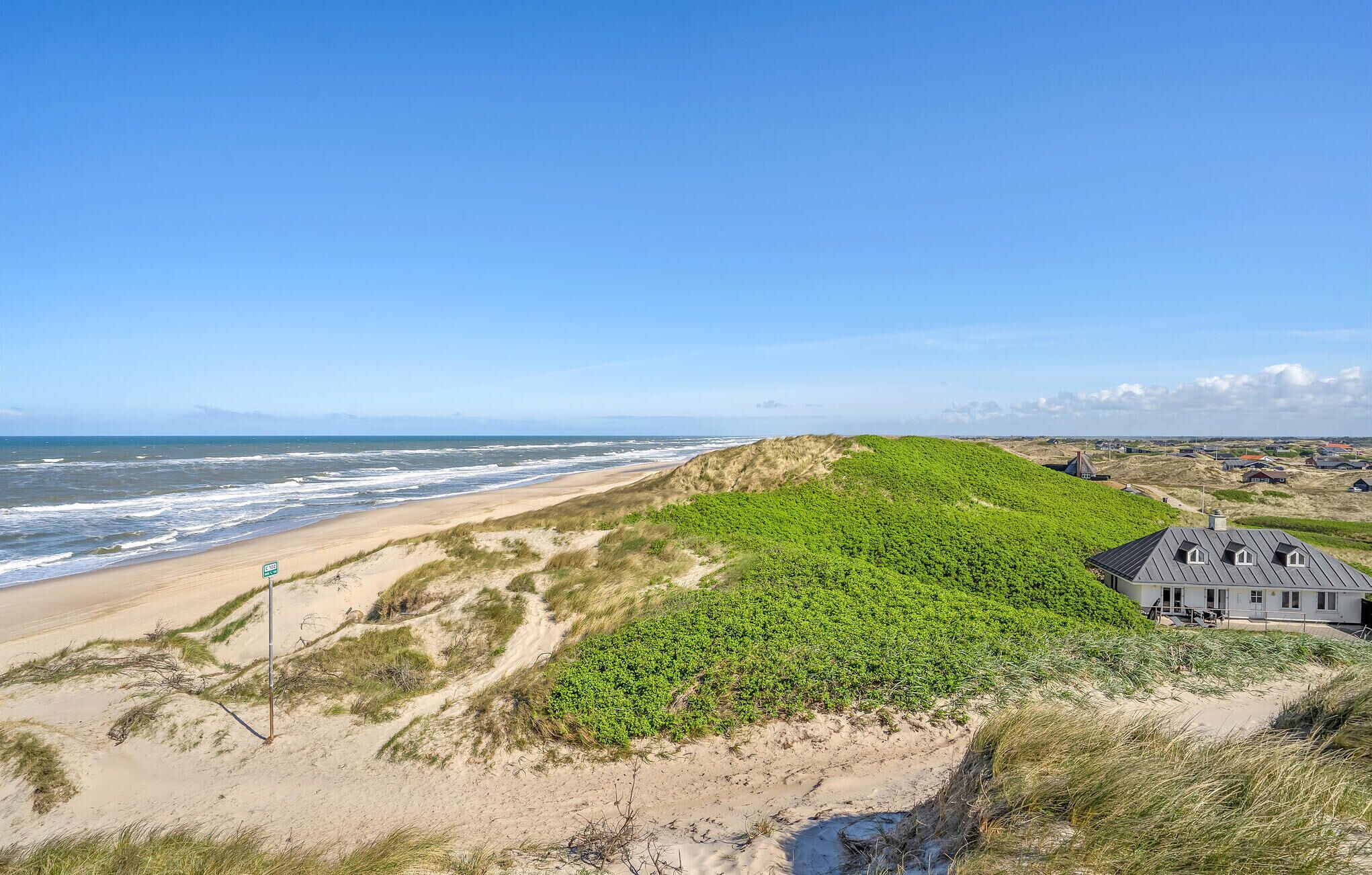Un paysage côtier avec une plage de sable, des dunes et un bâtiment doté d’un toit en pente.