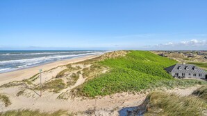 Un paysage côtier avec une plage de sable, des dunes et un bâtiment doté d’un toit en pente.