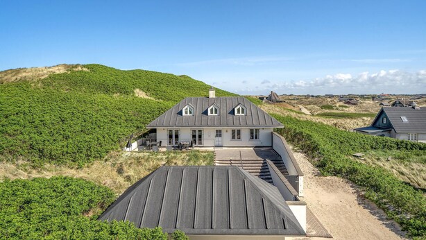 Une grande maison avec un toit en pente, entourée de collines verdoyantes et sous un ciel dégagé.