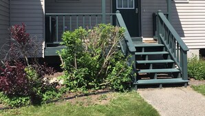 A two-story house with a green front porch, a front door with a wreath, and a small front yard with shrubs and a concrete path leading to the door.