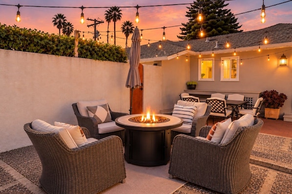 An outdoor patio area with wicker furniture, a central fire pit, string lights, and a view of the house.