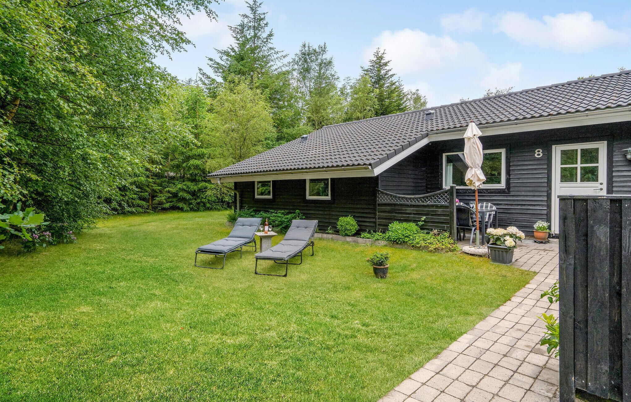 Une maison avec un toit en tuiles, un petit jardin avec de la pelouse, deux chaises longues et une clôture en bois.