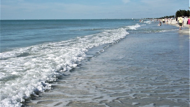 Une plage où les vagues viennent doucement lécher le rivage, un ciel dégagé et des gens qui se promènent le long de la plage.