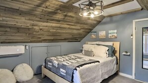 Un dormitorio con techo inclinado, vigas de madera, cama con manta estampada, almohadas mullidas y aire acondicionado montado en la pared.