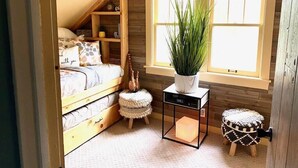 Un dormitorio acogedor con techo inclinado, una cama con cabecero de madera, una mesita de noche con una planta, un taburete pequeño y una mesita con una lámpara.