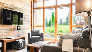 Ett modernt vardagsrum med träinredning, en platt-TV, en soffa, ett soffbord och ett stort fönster med utsikt över grönområden.