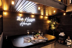 A bar area with a neon sign reading 'The Hygge House,' a wooden bar counter, glasses, a bottle, and a small plant.