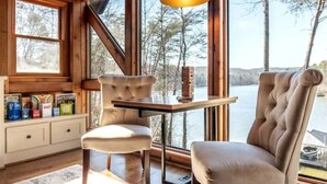 Una cabaña con un gran ventanal con vistas a un lago, una mesa de madera y un sillón con cojines.
