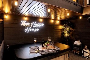 Zona de bar con un letrero de neón que dice "The Hygge House", una barra con vasos y botellas, y una planta decorativa.