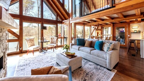 Amplia sala de estar con un sofá grande, una mesa de centro y una chimenea. Varias ventanas ofrecen luz natural y vistas al exterior.