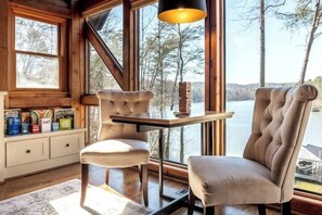 A room with a view of a lake, a wooden table, two chairs, and a shelf with books and containers.