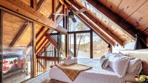 Un dormitorio con techo de madera, cama con ropa de cama blanca, un banco y ventana con vista a un lago.