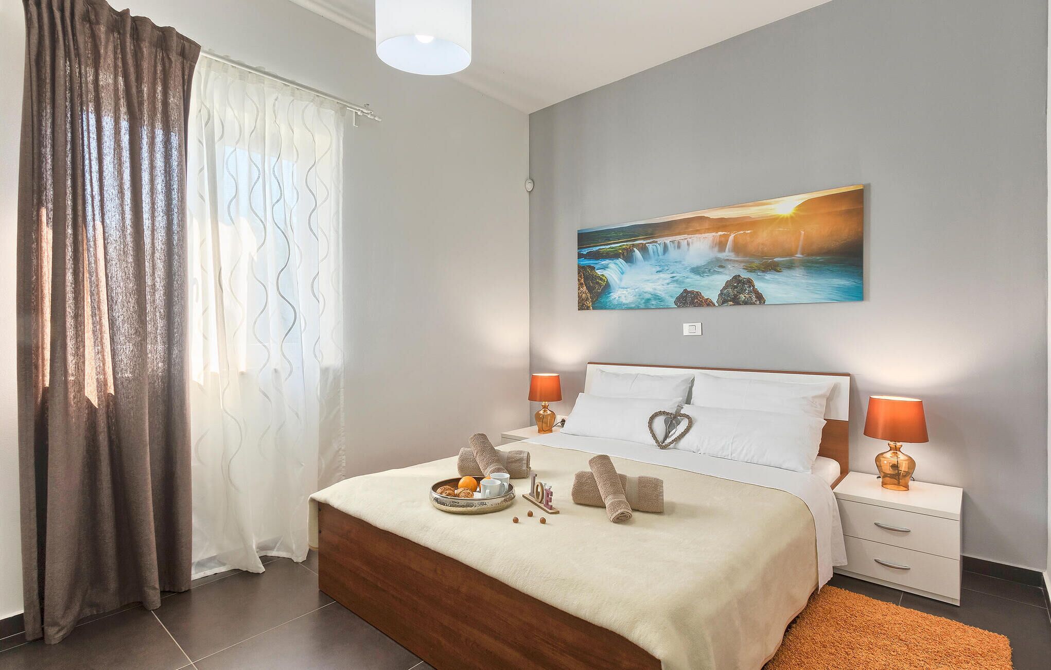 Une chambre à coucher avec un lit, deux tables de chevet avec des lampes, un grand tableau au mur et une fenêtre avec des rideaux.